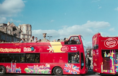 A brightly colored red double-decker bus with 'City Sightseeing Bath' written on the side. The bus features various vibrant designs, including the Union Jack and iconic city landmarks. People are seated on the upper open-top deck, enjoying the view of historical buildings and a clear sky.