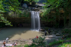 A serene waterfall cascades into a shallow pool surrounded by lush green foliage and rocky terrain. Several people are seen enjoying nature, including some near the water and others on the banks. The setting suggests a peaceful, outdoor gathering spot with a tent positioned near the top of the falls, indicating a camping area.