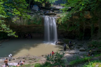 A serene waterfall cascades into a shallow pool surrounded by lush green foliage and rocky terrain. Several people are seen enjoying nature, including some near the water and others on the banks. The setting suggests a peaceful, outdoor gathering spot with a tent positioned near the top of the falls, indicating a camping area.