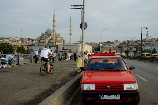 a red car on a road with people and a building in the background