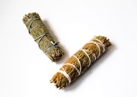 Two bundles of dried herbs bound with string rest against a white background. The herbs appear to be bound tightly, with one bundle wrapped in black string and the other in white string.