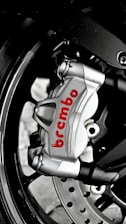 A close-up of a metallic brake caliper with the brand name 'Brembo' written in red. The caliper is attached to a motorcycle wheel, and features visible bolts and a disc rotor. The image highlights the engineering and design details of the brake system.