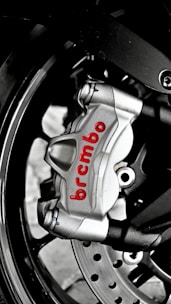 A close-up of a metallic brake caliper with the brand name 'Brembo' written in red. The caliper is attached to a motorcycle wheel, and features visible bolts and a disc rotor. The image highlights the engineering and design details of the brake system.