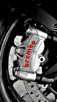 A close-up of a metallic brake caliper with the brand name 'Brembo' written in red. The caliper is attached to a motorcycle wheel, and features visible bolts and a disc rotor. The image highlights the engineering and design details of the brake system.