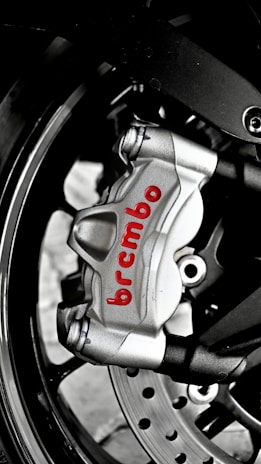 A close-up of a metallic brake caliper with the brand name 'Brembo' written in red. The caliper is attached to a motorcycle wheel, and features visible bolts and a disc rotor. The image highlights the engineering and design details of the brake system.