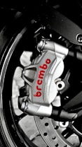 A close-up of a metallic brake caliper with the brand name 'Brembo' written in red. The caliper is attached to a motorcycle wheel, and features visible bolts and a disc rotor. The image highlights the engineering and design details of the brake system.