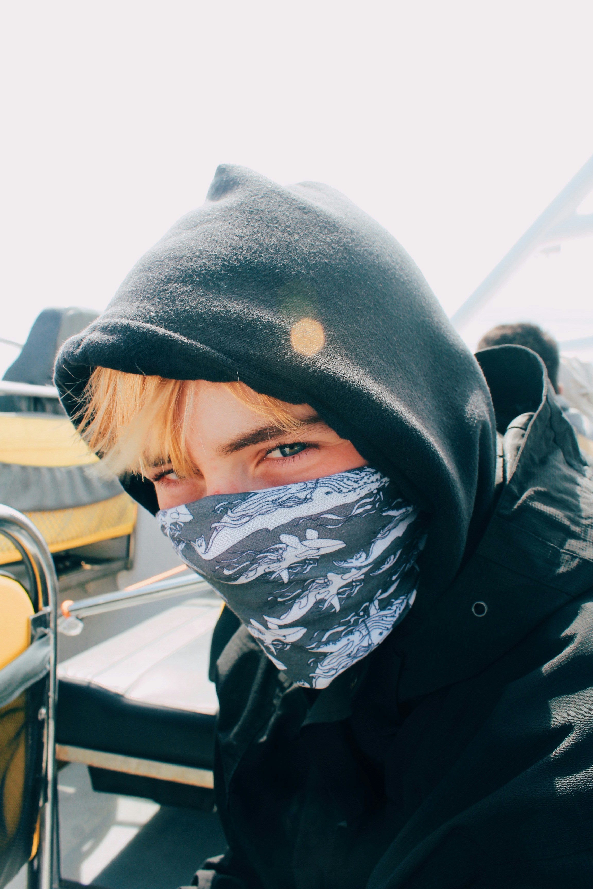 Young man with the hood of a dark sweatshirt up and a gray patterned gaiter pulled over his nose, sitting on a boat with a yellow cover behind him