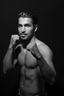 A black and white portrait of a shirtless, athletic man posing in a boxing stance with both fists raised in front of his face. The background is plain and dark, accentuating the subject's muscular physique and focused expression.