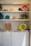 Shelves filled with various colored buckets, rustic tools, and plant pots. The bottom section features a wooden box holding magazines and a stack of papers next to more buckets.