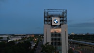 A tall building with a large BMW logo prominently displayed near the top, surrounded by a metal framework structure. The building is set against an evening sky with a mix of cityscape and greenery in the backdrop. A road with light traffic is visible in the foreground.