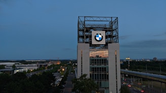 A tall building with a large BMW logo prominently displayed near the top, surrounded by a metal framework structure. The building is set against an evening sky with a mix of cityscape and greenery in the backdrop. A road with light traffic is visible in the foreground.