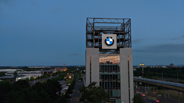 A tall building with a large BMW logo prominently displayed near the top, surrounded by a metal framework structure. The building is set against an evening sky with a mix of cityscape and greenery in the backdrop. A road with light traffic is visible in the foreground.