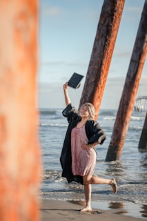 A person stands on a beach, posing under the shade of tall, rusted iron beams near the water. They are barefoot, wearing a black graduation gown over a pink dress, and holding a graduation cap in the air with a joyful expression. The ocean waves gently lap at the shore, and the sky is clear with light clouds.
