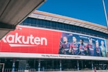 Large building facade featuring a prominent red Rakuten advertisement next to an image of six soccer players in team uniforms. The facade includes the slogan 'The Big Picture: Making history together'.