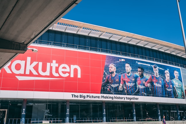 Large building facade featuring a prominent red Rakuten advertisement next to an image of six soccer players in team uniforms. The facade includes the slogan 'The Big Picture: Making history together'.