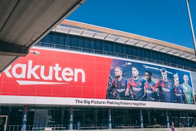 Large building facade featuring a prominent red Rakuten advertisement next to an image of six soccer players in team uniforms. The facade includes the slogan 'The Big Picture: Making history together'.