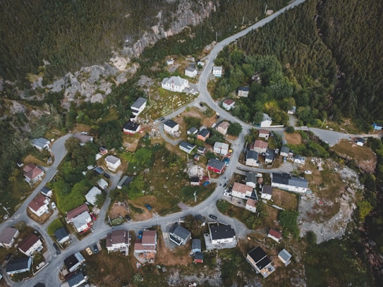 An aerial view of a small town nestled among lush greenery and rocky terrain, with winding roads connecting clusters of houses. The settlement features a variety of residential buildings, some painted in vibrant colors, set against a backdrop of dense forest. Vehicles are visible parked near the homes.