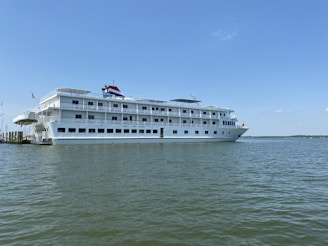 A large white cruise ship is docked on a calm body of water under a clear blue sky. The ship has multiple decks with rows of windows and external railings. An American flag is visible at the back of the vessel.