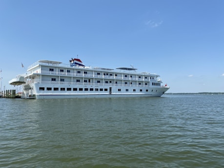 A large white cruise ship is docked on a calm body of water under a clear blue sky. The ship has multiple decks with rows of windows and external railings. An American flag is visible at the back of the vessel.