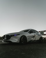 A sleek, sporty car with a white exterior and dark tinted windows is parked on a paved surface. The car's headlights have a distinctive bluish tint. It features custom graphics on the side and dark wheels. In the background, there is a partially visible caution sign indicating development on the street.