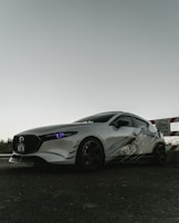 A sleek, sporty car with a white exterior and dark tinted windows is parked on a paved surface. The car's headlights have a distinctive bluish tint. It features custom graphics on the side and dark wheels. In the background, there is a partially visible caution sign indicating development on the street.
