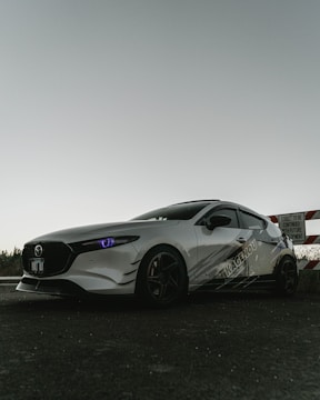 A sleek, sporty car with a white exterior and dark tinted windows is parked on a paved surface. The car's headlights have a distinctive bluish tint. It features custom graphics on the side and dark wheels. In the background, there is a partially visible caution sign indicating development on the street.