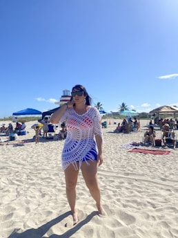 A person stands barefoot on a sandy beach, wearing sunglasses and a crocheted white cover-up over a blue outfit. In the background, people are relaxing under beach umbrellas. The sky is clear with a few small clouds, and there are palm trees and a structure resembling a lighthouse.