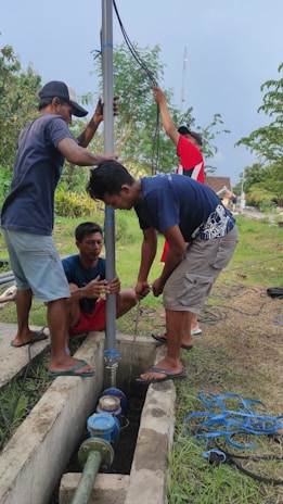 A group of four men are working together to install or repair a vertical pipe outdoors. They are holding and adjusting the pipe, which is situated in a narrow concrete trench with some type of machinery at the base. The surrounding environment includes grassy areas, some bushes, and trees. There are cables lying around on the grass, and it appears to be a somewhat overcast day.