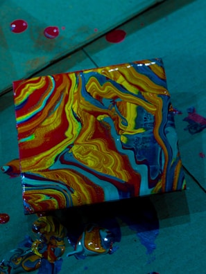 A square canvas features swirling, vibrant patterns of red, yellow, blue, and orange paint. The paint appears to have been poured and allowed to flow naturally, creating a marbled effect. Surrounding the canvas are small splatters and blobs of paint, indicating a spontaneous and dynamic art-making process. The background is a muted, shadowy teal surface.