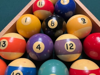 A triangular arrangement of billiard balls on a green surface, featuring a variety of colors such as red, yellow, blue, purple, and black. Each ball is marked with a number, ranging from 1 to 15. A wooden rack secures the balls in place.