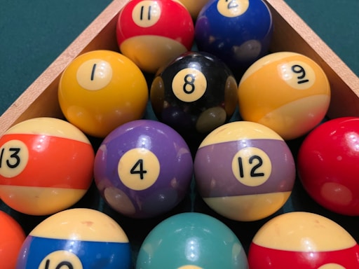 A triangular arrangement of billiard balls on a green surface, featuring a variety of colors such as red, yellow, blue, purple, and black. Each ball is marked with a number, ranging from 1 to 15. A wooden rack secures the balls in place.