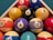 A triangular arrangement of billiard balls on a green surface, featuring a variety of colors such as red, yellow, blue, purple, and black. Each ball is marked with a number, ranging from 1 to 15. A wooden rack secures the balls in place.