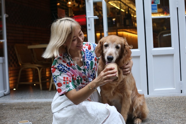 A person with blonde hair wearing a colorful floral top and white skirt is sitting with a golden retriever. They are offering the dog a cup, as they both sit near the entrance of a café or restaurant. The setting is cozy, with warm lighting coming from inside.