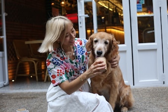 A person with blonde hair wearing a colorful floral top and white skirt is sitting with a golden retriever. They are offering the dog a cup, as they both sit near the entrance of a caf&eacute; or restaurant. The setting is cozy, with warm lighting coming from inside.