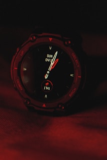 A digital wristwatch displaying the time and date, with the background illuminated in deep red lighting. The watch's face includes traditional clock hands and digital indicators, surrounded by a rugged, dark casing.