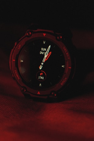 A digital wristwatch displaying the time and date, with the background illuminated in deep red lighting. The watch's face includes traditional clock hands and digital indicators, surrounded by a rugged, dark casing.