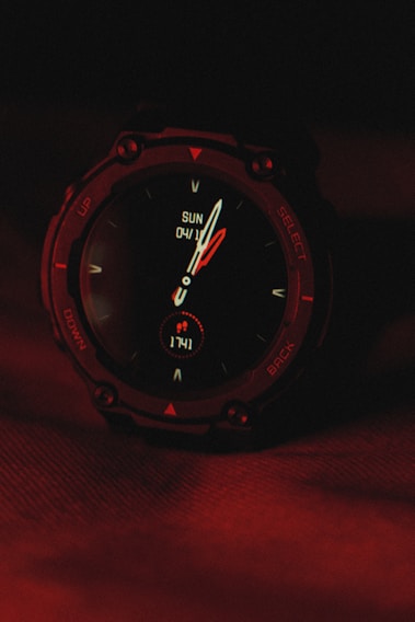 A digital wristwatch displaying the time and date, with the background illuminated in deep red lighting. The watch's face includes traditional clock hands and digital indicators, surrounded by a rugged, dark casing.