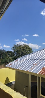 A metal-roofed building is situated in the foreground, showing signs of rust. The building is painted yellow and stands next to a concrete wall. Behind the building, a tree is visible, and the background consists of a clear blue sky with scattered white clouds.