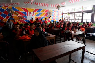 A classroom filled with students wearing matching red and black uniforms. They sit at wooden desks, facing forward, with a few students engaging in conversation. The room has a colorful geometric-patterned wall that displays 'IPA 3' in bold letters. Several small potted plants hang from the ceiling and around the windows, adding greenery to the space. Light streams in through large windows on one side, illuminating the students.