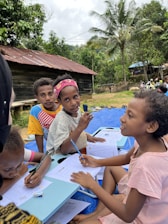 Several children are sitting outdoors on a blue mat, engaging in writing activities. They have papers and pencils in their hands, appearing focused and attentive. The background features lush green trees, a rustic wooden house with a tin roof, and some people in the distance, suggesting a rural setting.