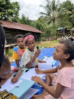 Several children are sitting outdoors on a blue mat, engaging in writing activities. They have papers and pencils in their hands, appearing focused and attentive. The background features lush green trees, a rustic wooden house with a tin roof, and some people in the distance, suggesting a rural setting.