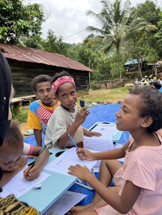 Several children are sitting outdoors on a blue mat, engaging in writing activities. They have papers and pencils in their hands, appearing focused and attentive. The background features lush green trees, a rustic wooden house with a tin roof, and some people in the distance, suggesting a rural setting.