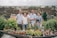 A family of six stands together in front of a scenic backdrop. There are various potted plants and flowers on a table in the foreground. Behind them, there is a view of rooftops and greenery, suggesting an elevated or rooftop garden setting. Everyone is smiling, conveying a sense of togetherness and happiness.