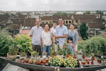 A family of six stands together in front of a scenic backdrop. There are various potted plants and flowers on a table in the foreground. Behind them, there is a view of rooftops and greenery, suggesting an elevated or rooftop garden setting. Everyone is smiling, conveying a sense of togetherness and happiness.