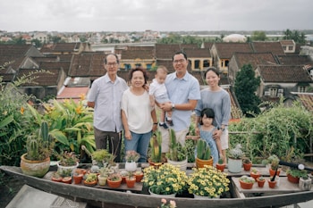 A family of six stands together in front of a scenic backdrop. There are various potted plants and flowers on a table in the foreground. Behind them, there is a view of rooftops and greenery, suggesting an elevated or rooftop garden setting. Everyone is smiling, conveying a sense of togetherness and happiness.