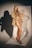 A woman with long blonde hair is posing confidently in a studio setting. She is wearing a beige blazer and high-cut nude bodysuit, paired with black high heels. The background displays a strong shadow created by focused lighting.