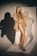 A woman with long blonde hair is posing confidently in a studio setting. She is wearing a beige blazer and high-cut nude bodysuit, paired with black high heels. The background displays a strong shadow created by focused lighting.