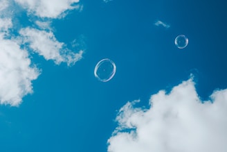 Two transparent soap bubbles floating against a backdrop of a clear blue sky scattered with a few fluffy white clouds.