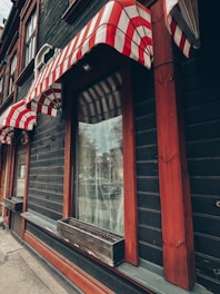 A building facade featuring striped red and white awnings over large windows. The structure has dark wood paneling with accents of red trim. Each window has an empty wooden planter box affixed beneath it. The street can be faintly seen reflected in the windows.