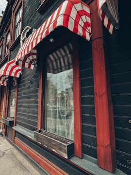 A building facade featuring striped red and white awnings over large windows. The structure has dark wood paneling with accents of red trim. Each window has an empty wooden planter box affixed beneath it. The street can be faintly seen reflected in the windows.